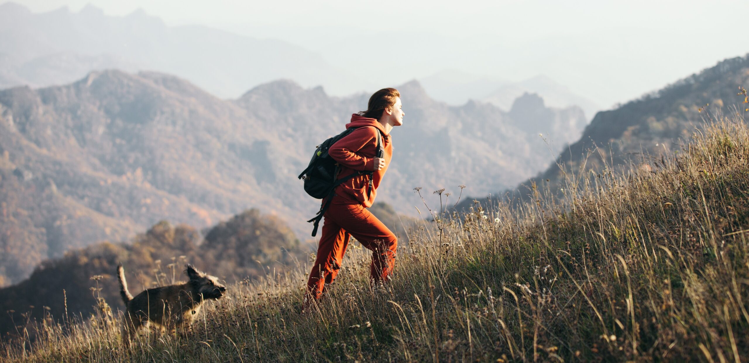 Beautiful woman traveler climbs uphill with a dog on a background of mountain views. She is with a backpack and in red clothes.